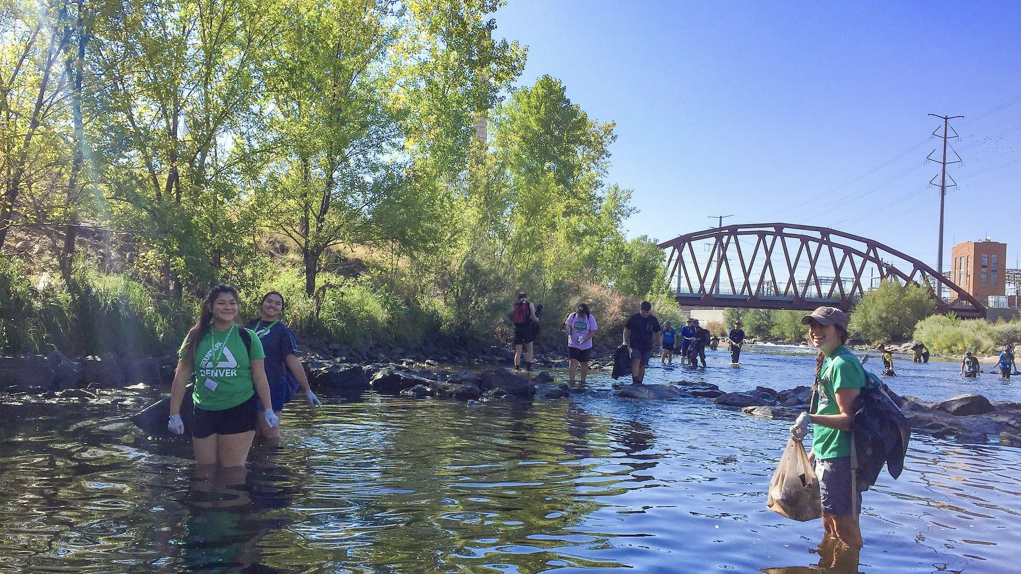 two smiling girls in green shirts holding bags of trash while standing in the middle of a river.