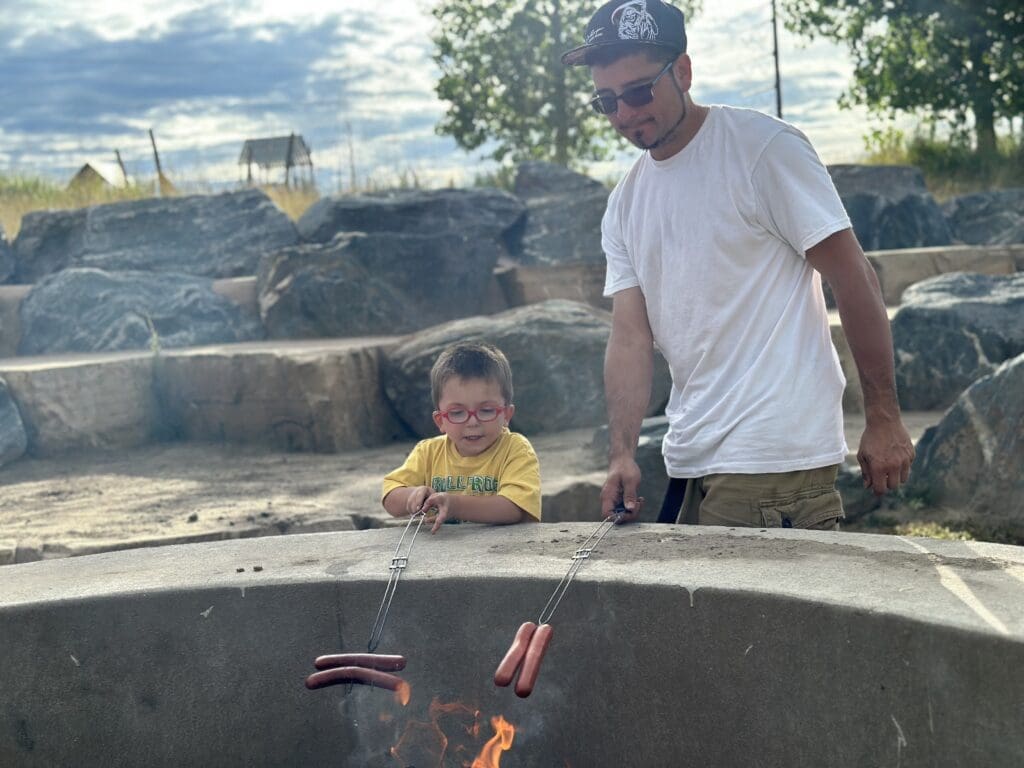 A Dad in a white shirt and black hat and son in red glasses and yellow shirt roast hot dogs over the fire.